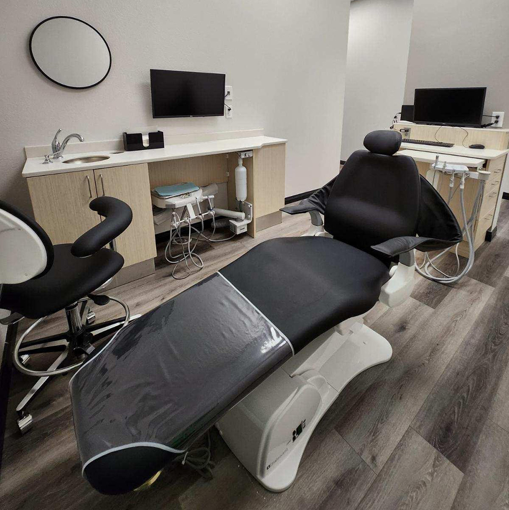The image shows an interior view of a dental office with a modern dental chair, a small counter with a sink and a mirror, and a television screen on the wall.